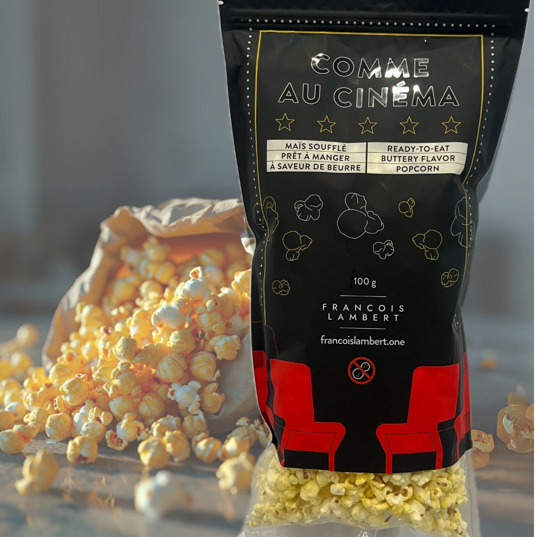 La photo montre un paquet de pop-corn prêt à manger avec la mention "COMME AU CINÉMA". Le paquet est noir avec des motifs de pop-corn et des étoiles dorées, indiquant une saveur de beurre. Il y a 100g de produit et le nom "FRANCOIS LAMBERT" est visible, avec un site web indiqué : francoislambert.one. À l'avant-plan, du pop-corn semble avoir été versé d'une portion du paquet, suggérant que le produit est directement consommable.