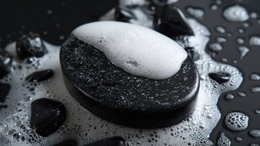 Un savon ovale noir posé sur un amas de gros graviers, avec un fond entièrement noir. L'arrière-plan présente de grandes pierres, de la fumée ainsi que des jeux de lumière et d'ombre.