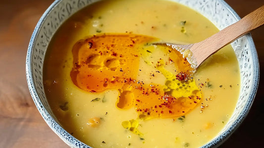 Une photo d'une soupe aux pois chiches crémeuse et jaune, servie dans une assiette en céramique blanche. La soupe est garnie d'une cuillère en bois et d'un filet d'huile piquante.