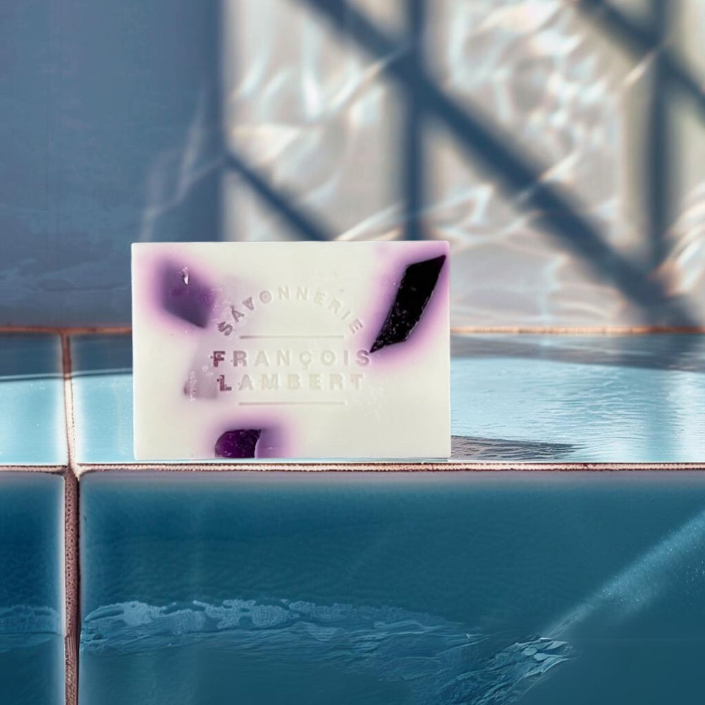 Savon au lait de brebis et trois beurres à la fragrance de raisin et bleuet posé au bord d'une piscine carrelée, avec des reflets d'eau en arrière-plan.